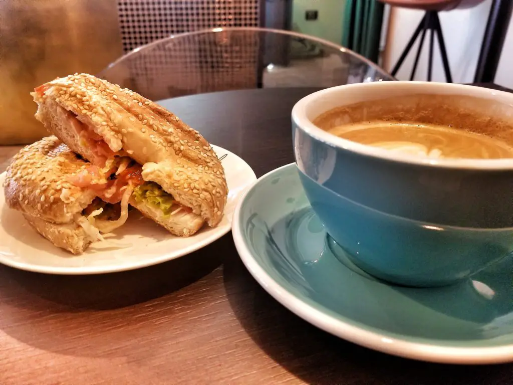 Flat white in turquoise mug alongside a prosciutto and tomato bagel sandwich.