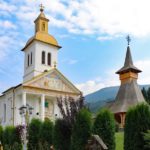 Two wooden churches in Maramures, with mountains in the background and green, lush bushes in the foreground under blue and white skies.