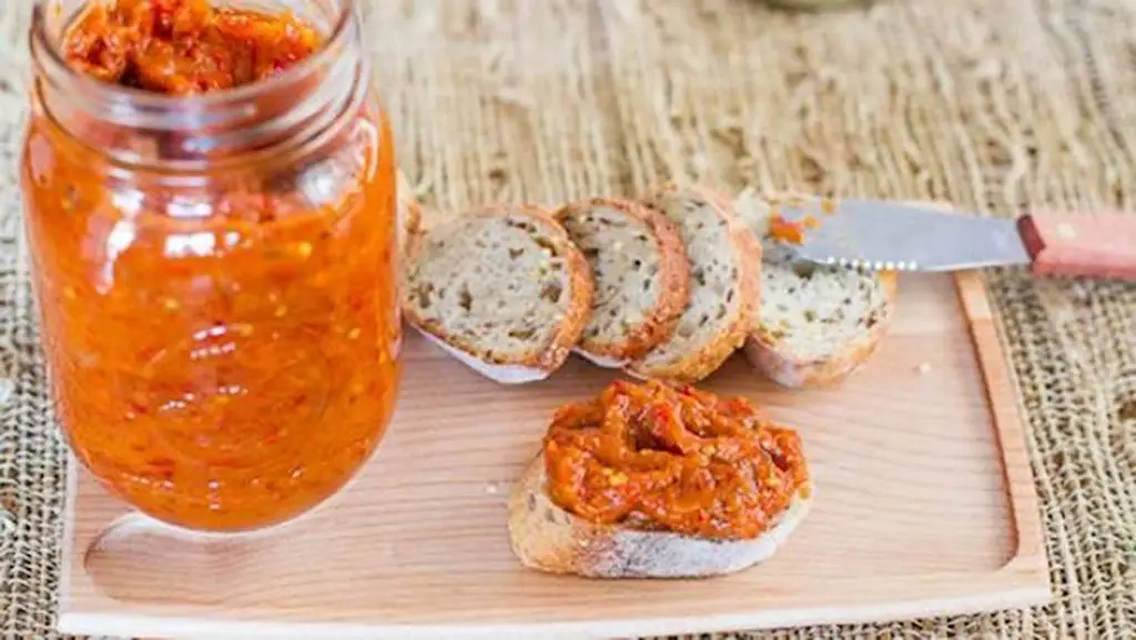 Jar of zacusca, alongside slices of bread, with one slice of bread topped with the orange zacusca.