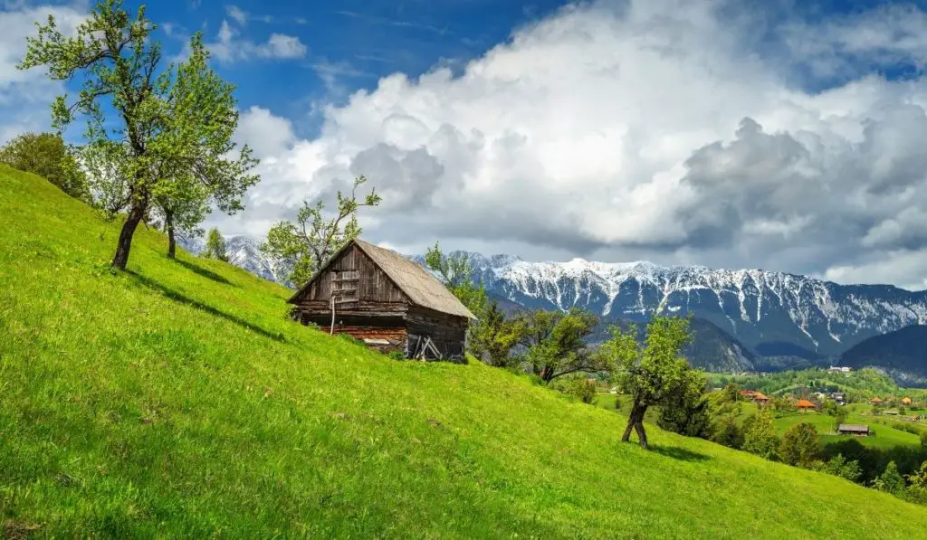 Transylvania landscape with wooden cottage on a hill in the mountains with lush green grass and cloudy skies.