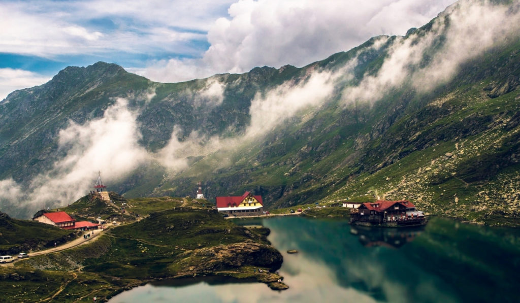 Balea Lake with low-lying clouds hanging around the mountains and a few scattered red-roofed houses on the shore of the lake.