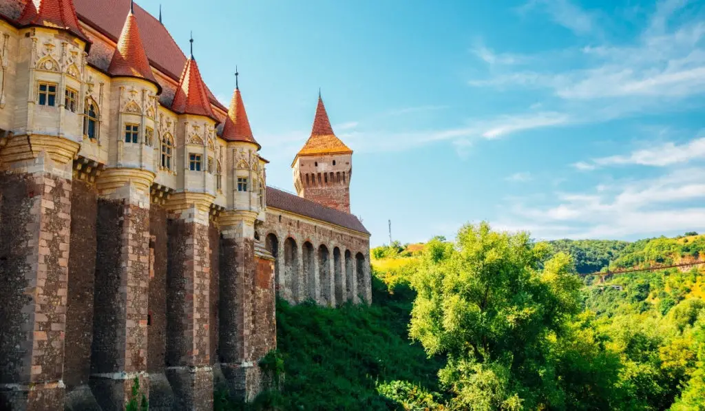 Outer wall of Hunedoara Castle with red spired towers and lush green countryside to the right.