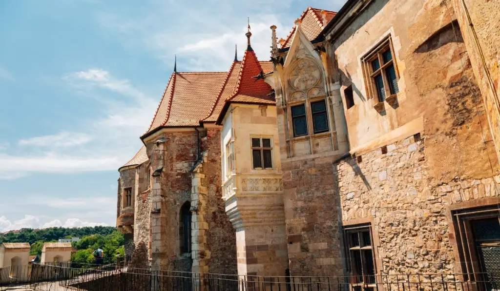 Exterior of Corvin Castle in Hunedoara, Romania with rectangular and circular towers, red roofs, and stone facade.