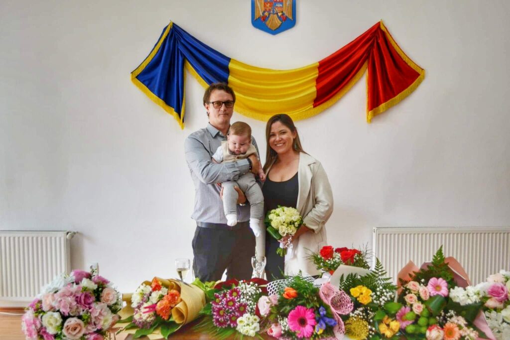 Romanian civil wedding ceremony - couple in front of Romanian flag and behind many bouquets of flowers.